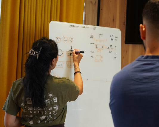 Person writing on a whiteboard with another person observing, against a yellow curtain.