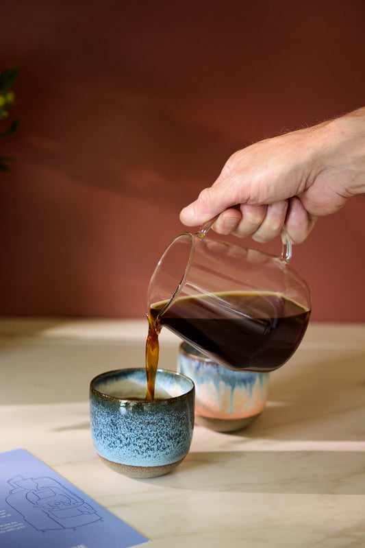 Hand pouring coffee from a glass pitcher into a small blue ceramic cup on a wooden surface.