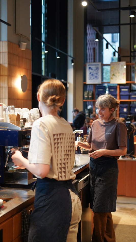 Two women in a coffee shop, one behind the counter and the other interacting.